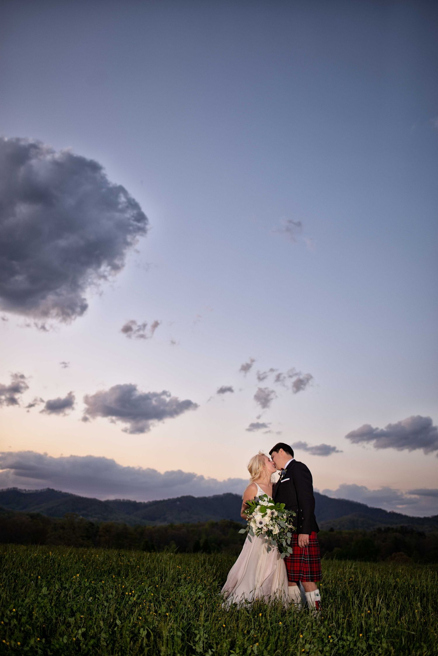 Bride and groom kissing in a green field at sunset, groom wearing a red tartan kilt, mountains in the background.