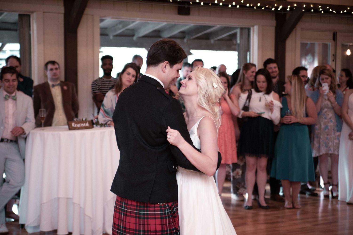 Bride and groom share first dance at wedding reception, groom wearing traditional red Scottish kilt, guests applauding in background.