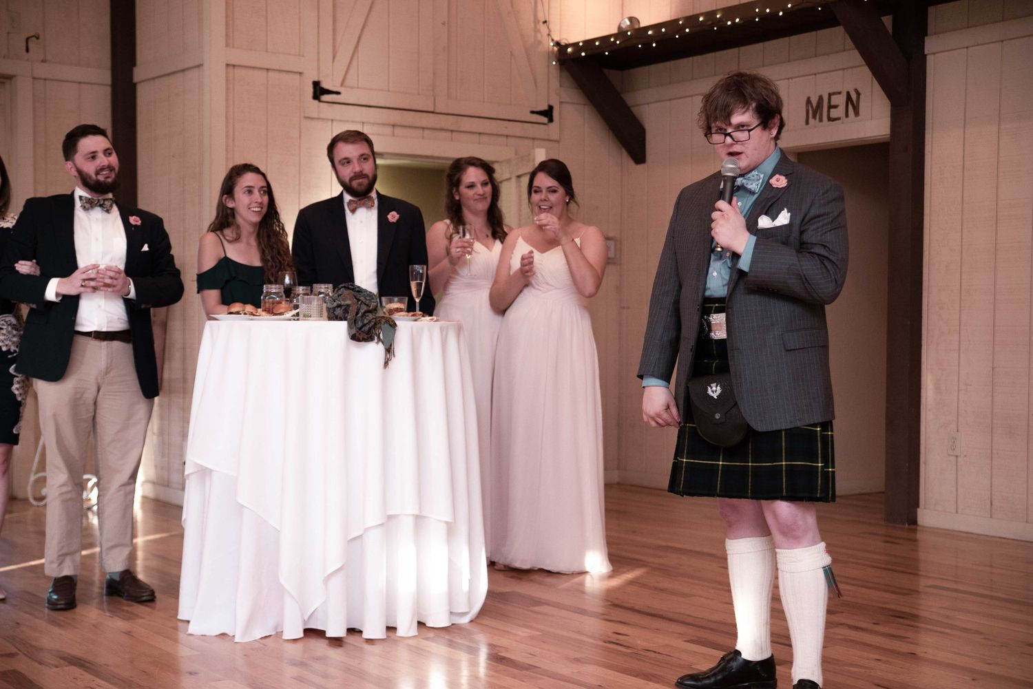 Man in kilt giving speech at wedding reception with bridal party standing near decorated table indoors.