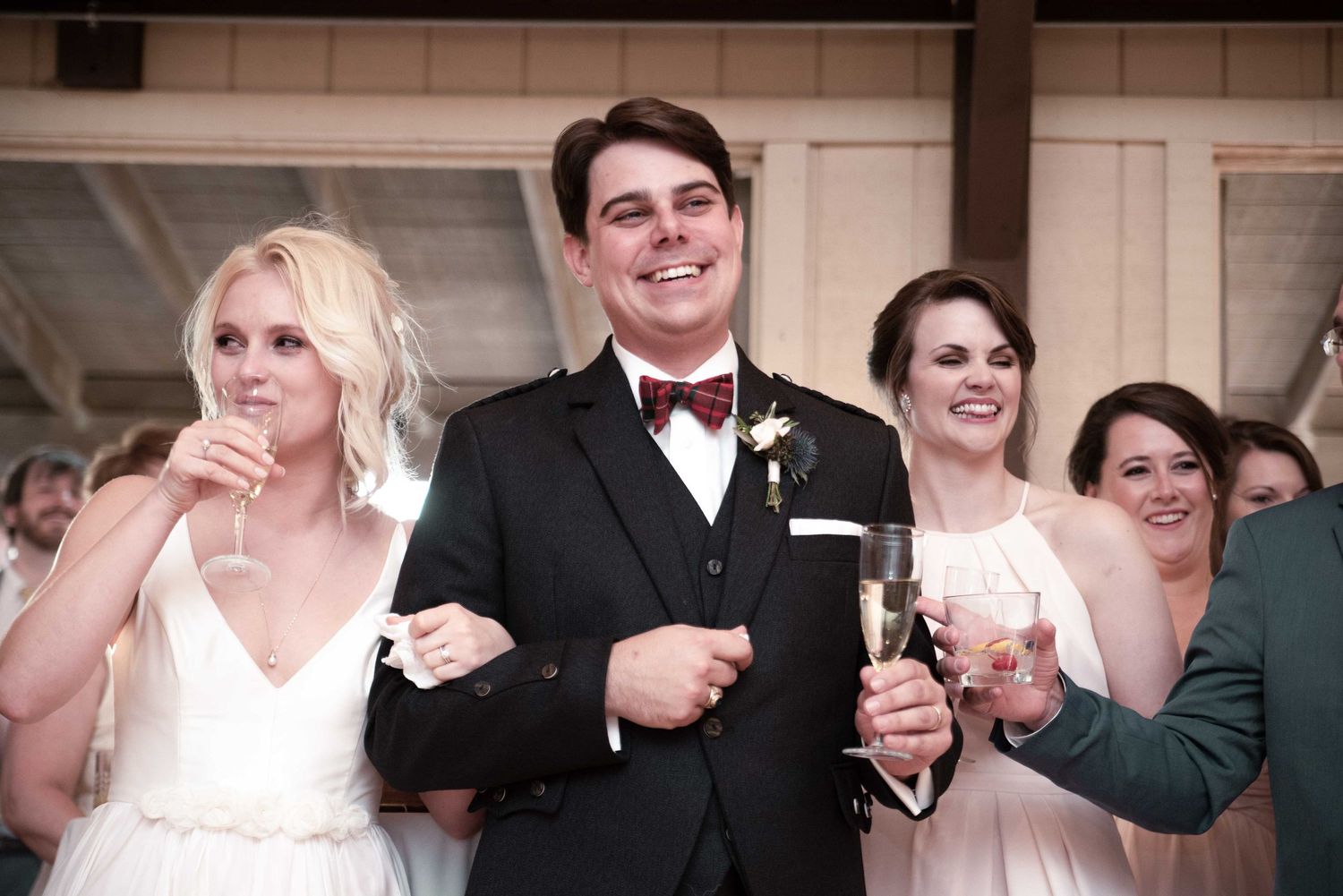 Groom in black tuxedo with bow tie raises champagne glass with bridesmaids in pink dresses at wedding reception.