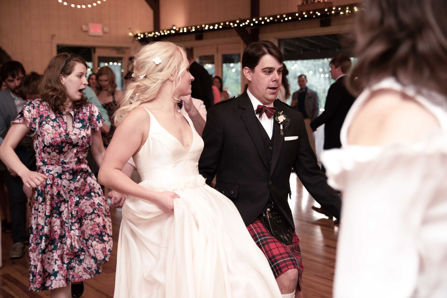 Bride and groom dancing joyfully at wedding reception, groom wearing kilt and bow tie, guests celebrating in background.