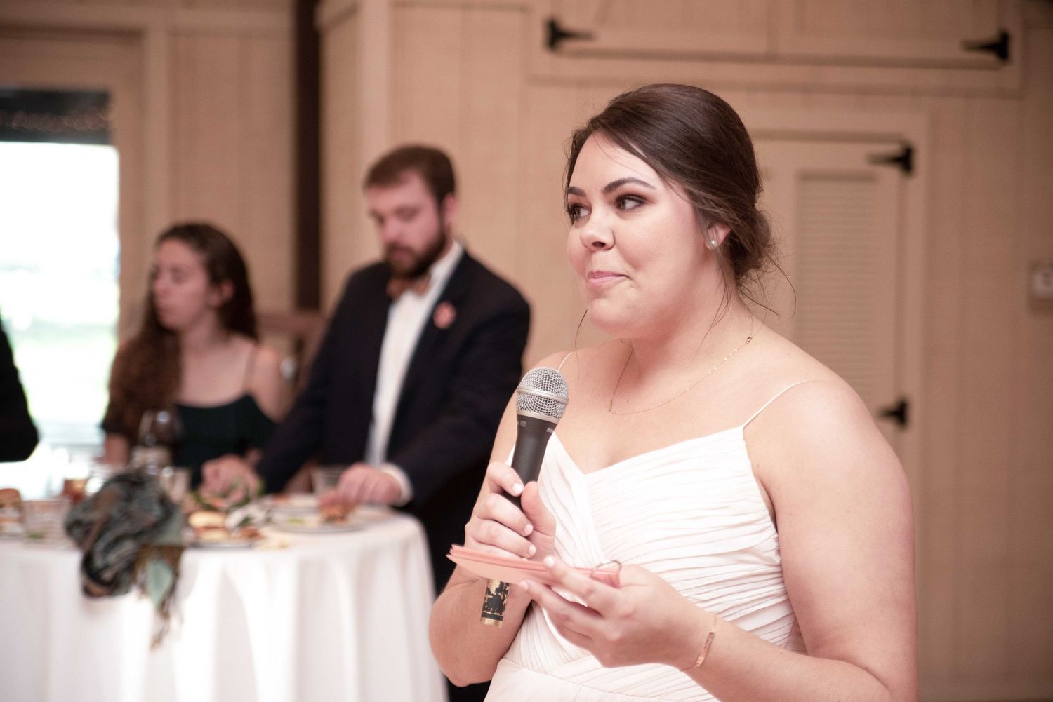 Bride in white strapless gown speaking into microphone during wedding reception speech.