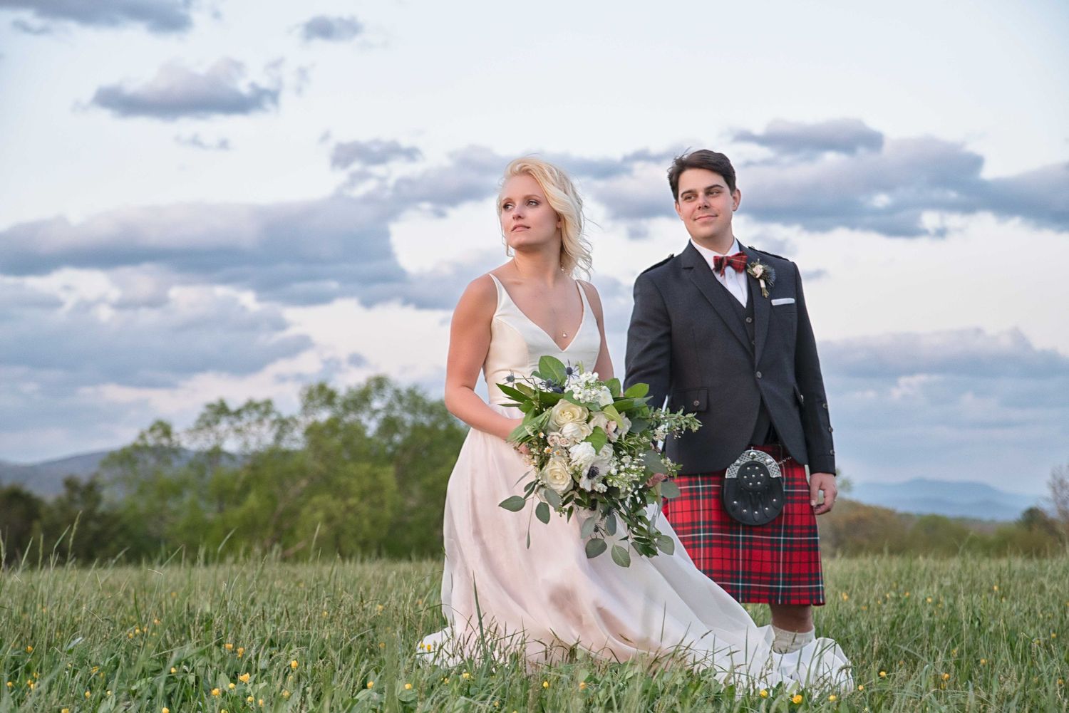 Bride in white gown and groom in tartan kilt pose in a lush green field with mountain backdrop.