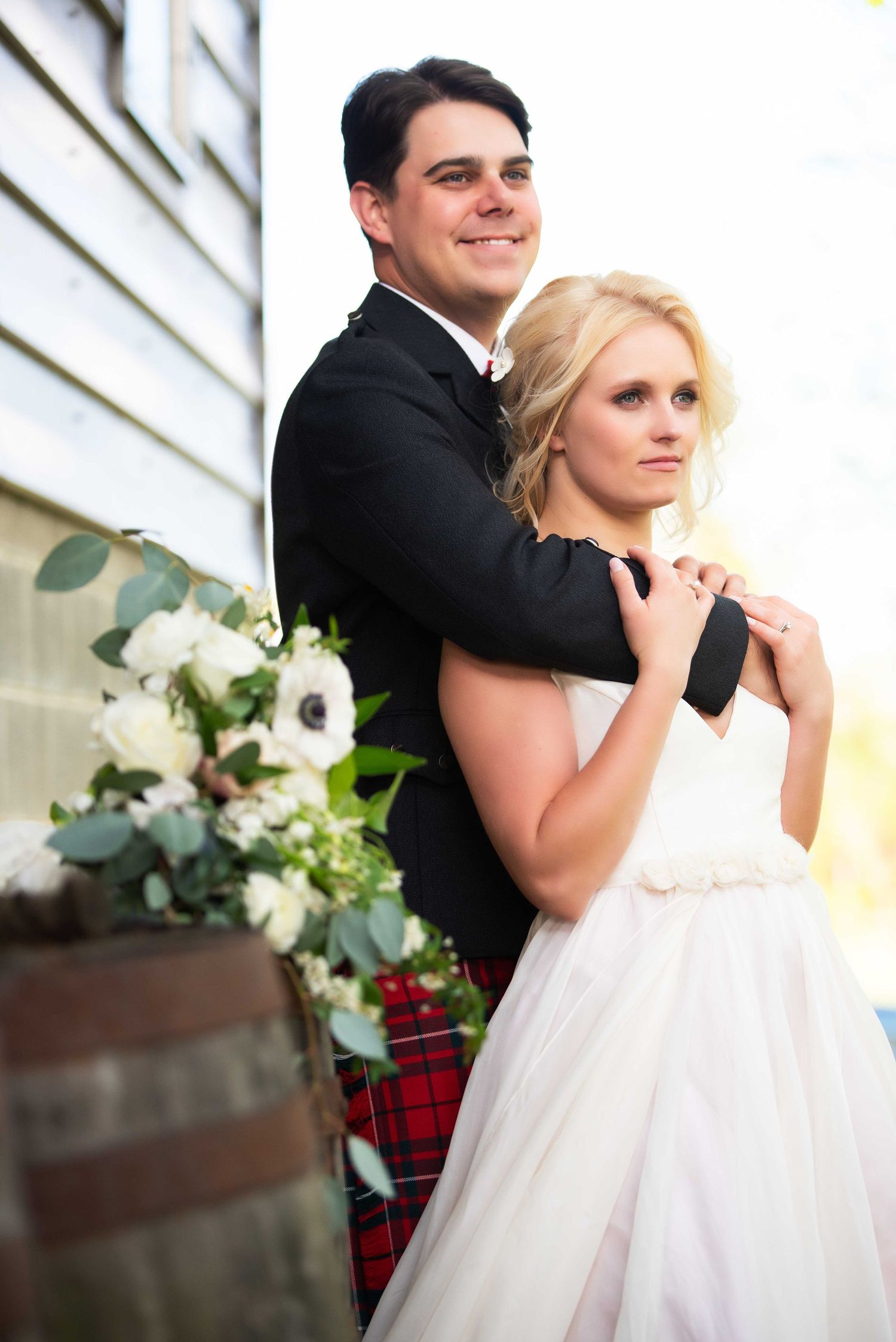 Bride and groom portrait outdoors, groom in tartan kilt embracing blonde bride holding white floral bouquet.
