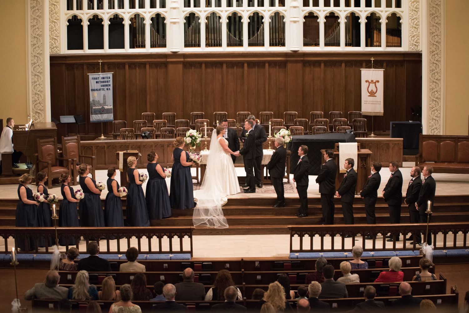 Wedding ceremony in a grand church with bridesmaids in navy dresses and groomsmen flanking the altar.