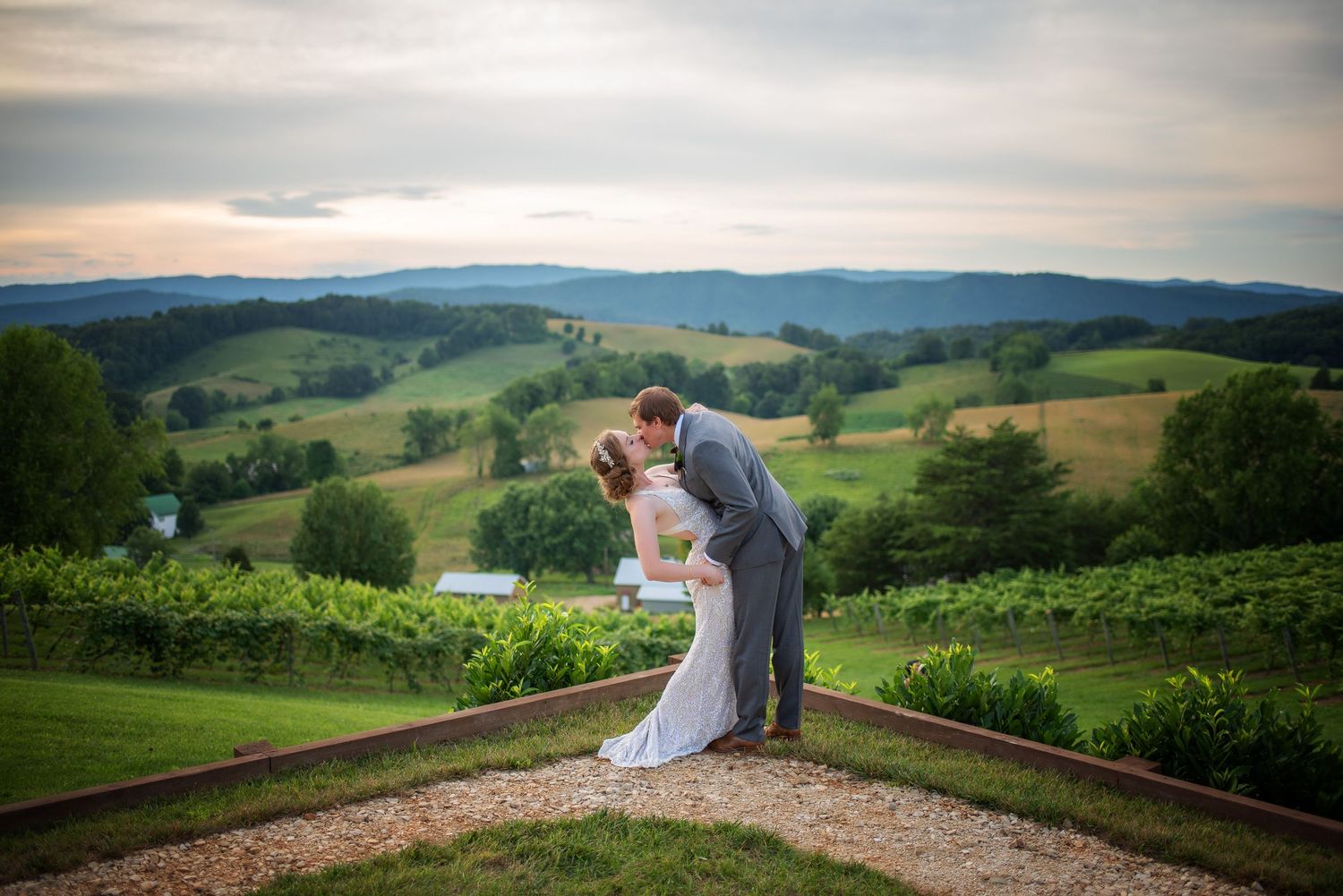 Bride and groom share a romantic dip kiss overlooking lush green vineyard hills at sunset.