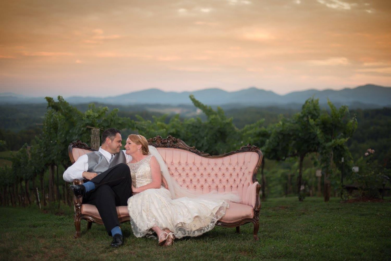 Bride and groom sitting on a pink vintage sofa in a vineyard at sunset with mountain views.
