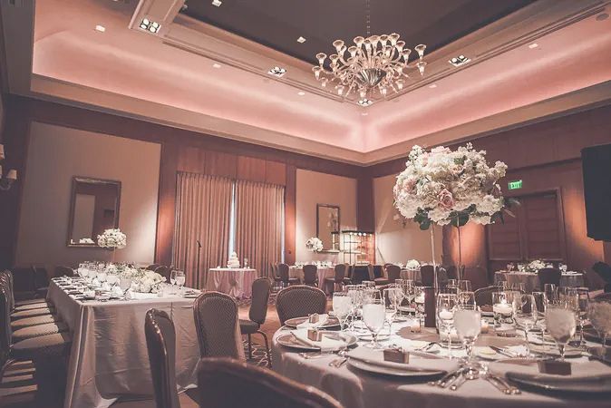 Elegant wedding reception ballroom with tall floral centerpieces, chandelier, and soft pink lighting.