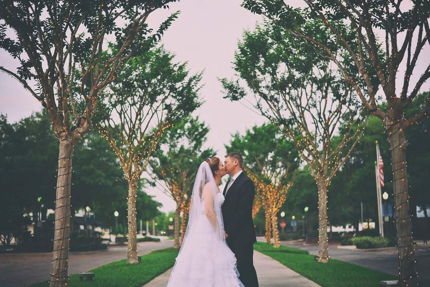 Newlyweds share a romantic kiss along a tree-lined pathway adorned with twinkling lights.