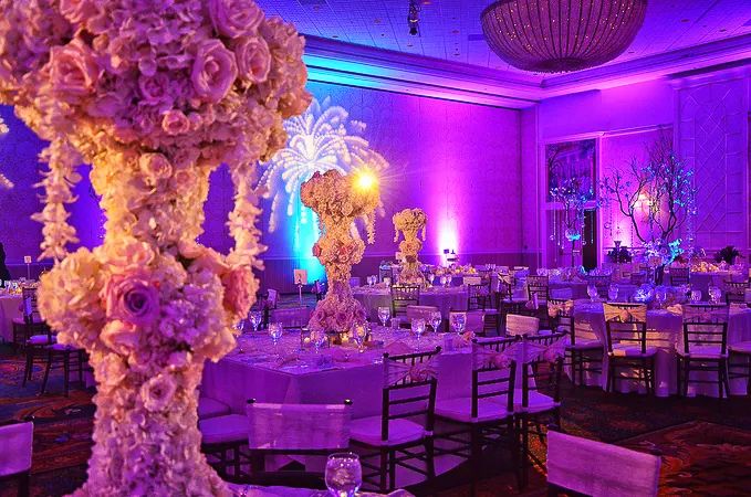 Elegant wedding reception hall with pink floral centerpieces, purple uplighting, and round tables with chiavari chairs.