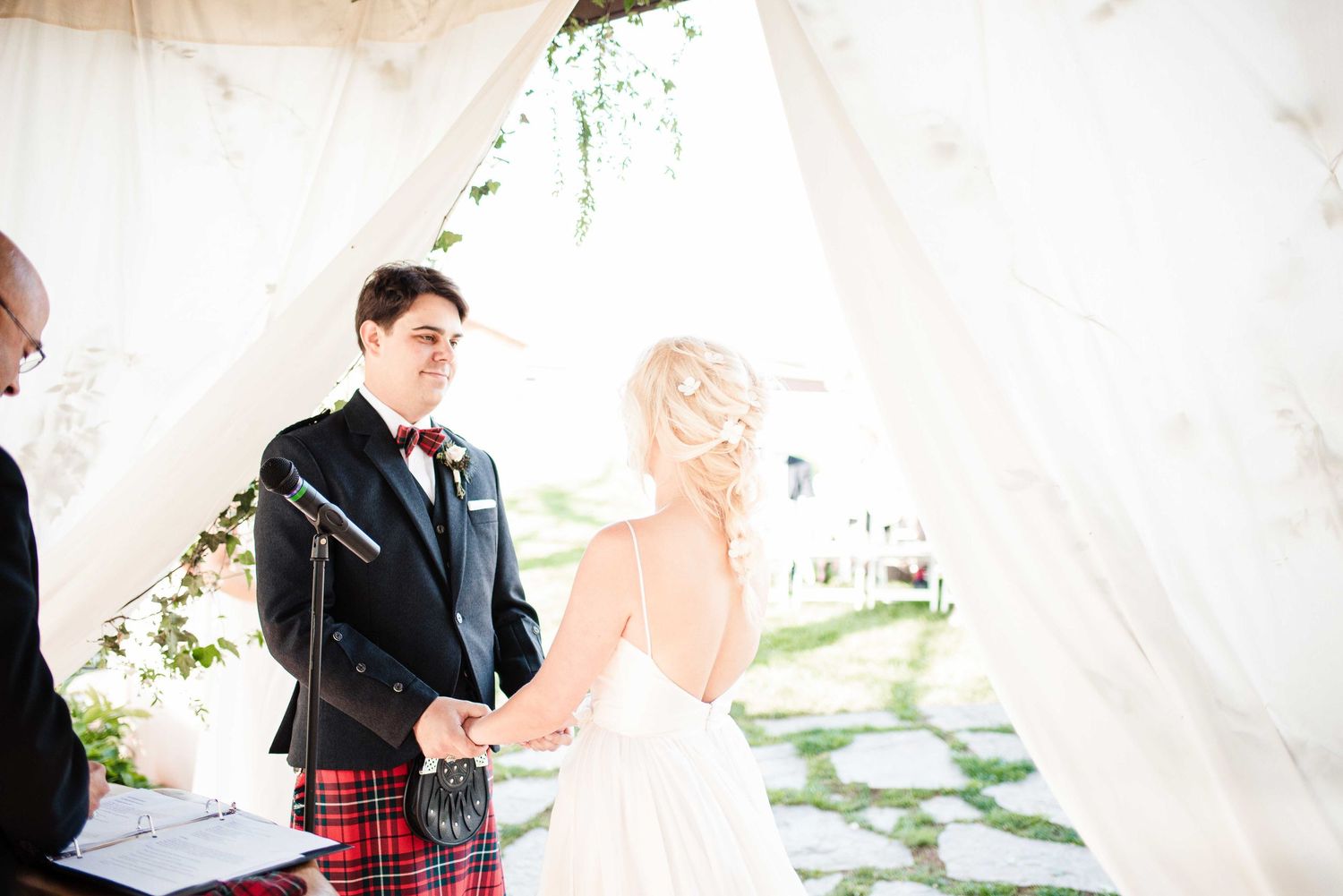 Bride and groom holding hands at outdoor wedding ceremony, groom wearing traditional Scottish tartan kilt and black jacket.