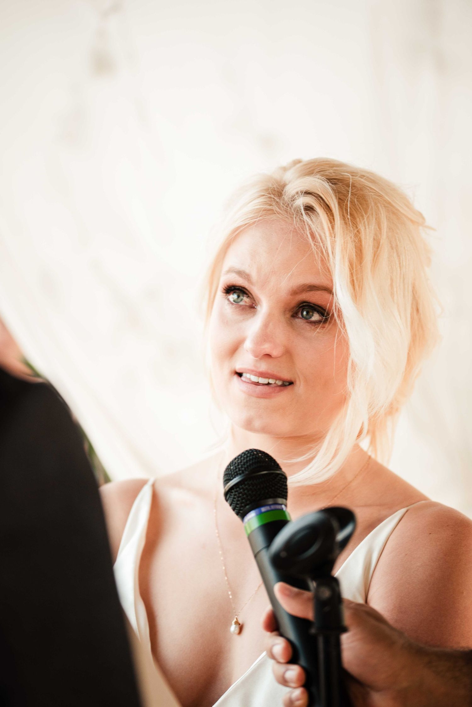 Blonde woman speaking into a microphone at a wedding reception, looking up with a warm smile.