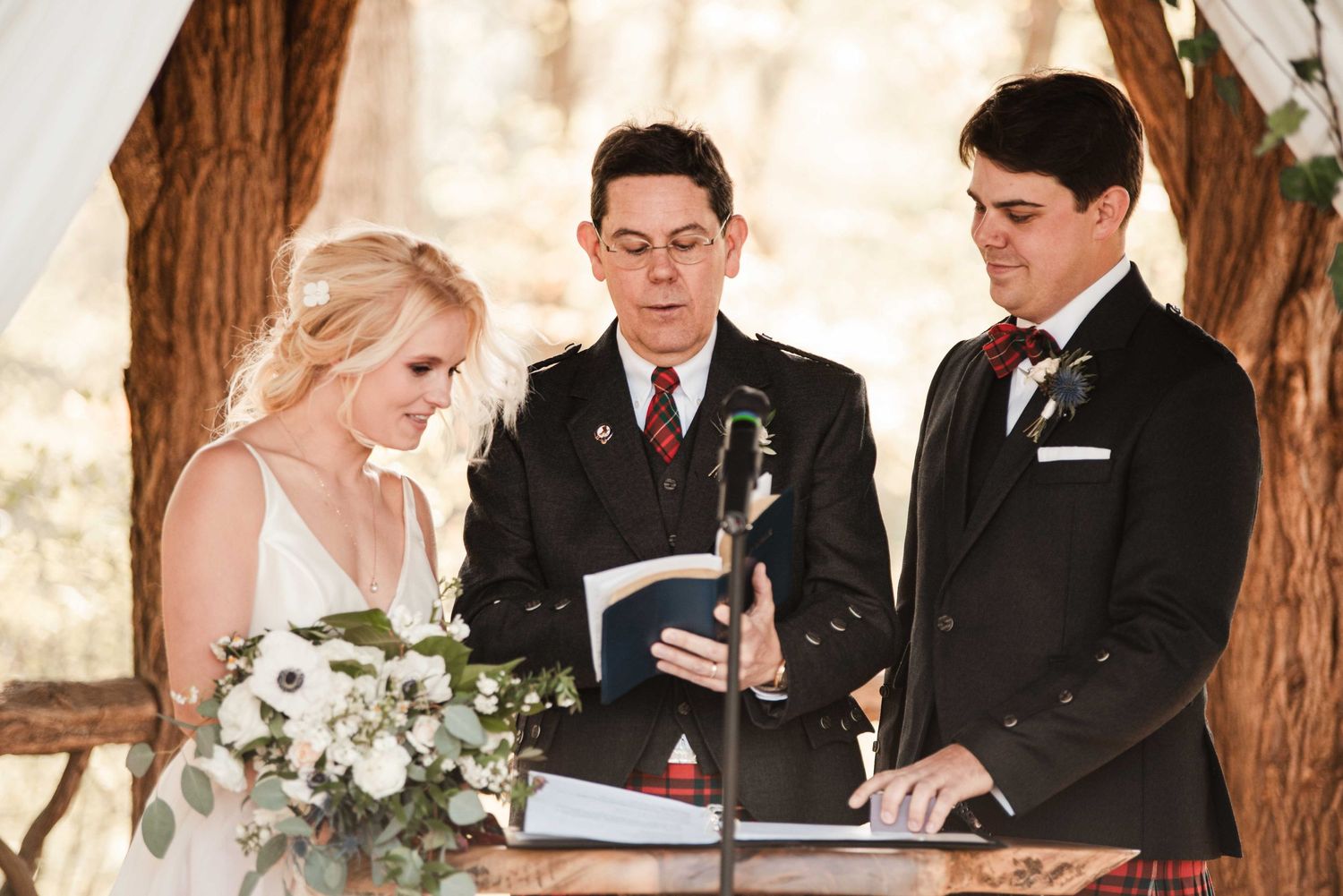 Bride and groom exchange vows with officiant at outdoor woodland wedding ceremony under rustic arch.