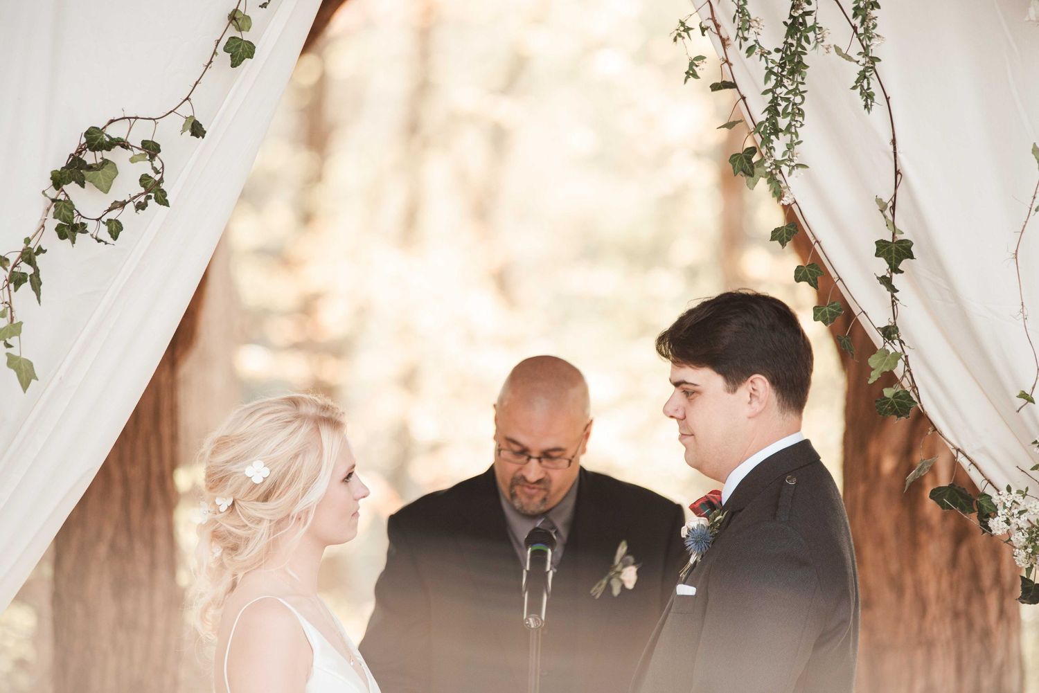 Bride and groom exchange vows under a floral arch with white drapes, officiated by a bald man at an outdoor wedding.