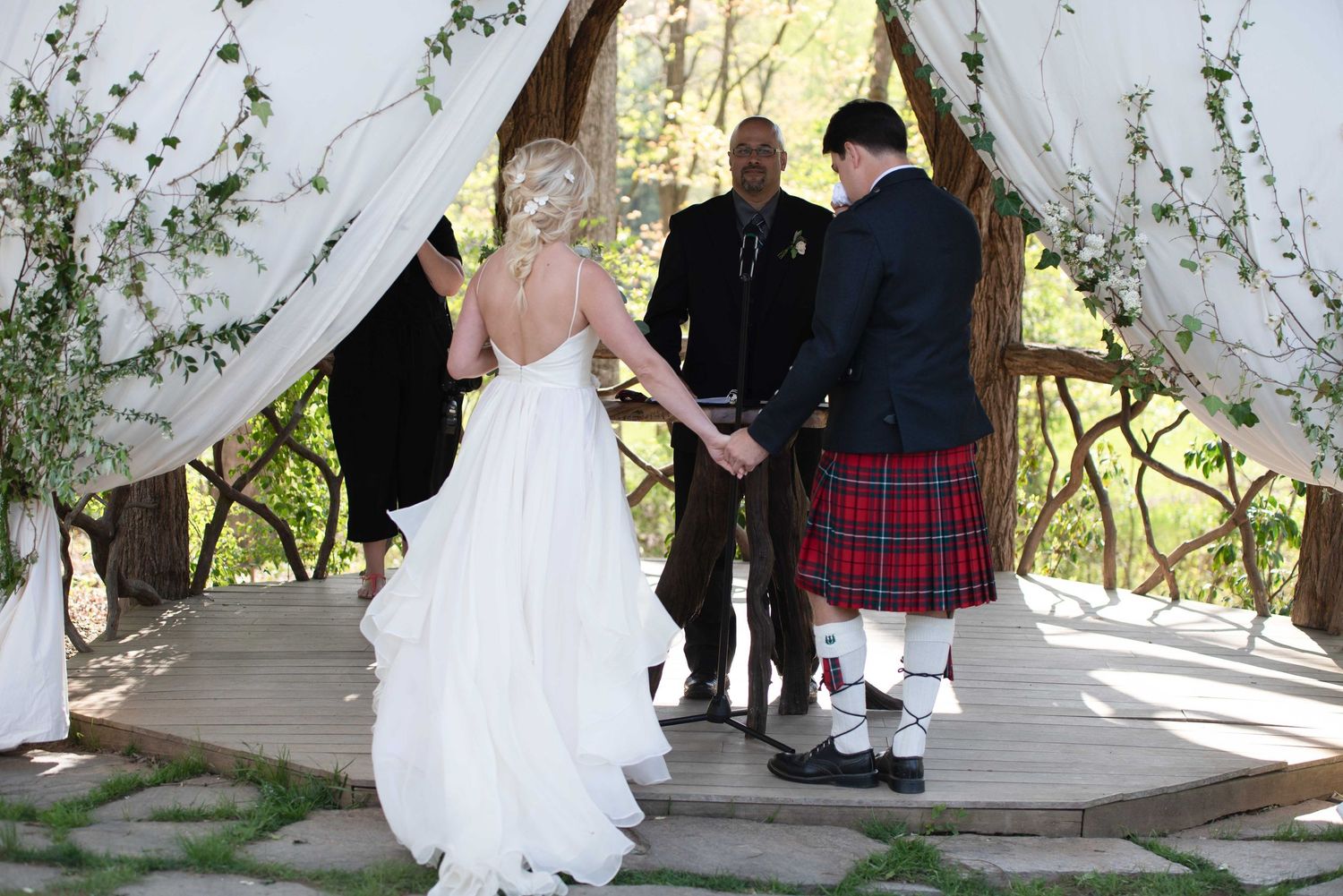Bride and groom holding hands at outdoor wedding ceremony, groom wearing red tartan kilt, officiant standing beneath rustic floral arch.