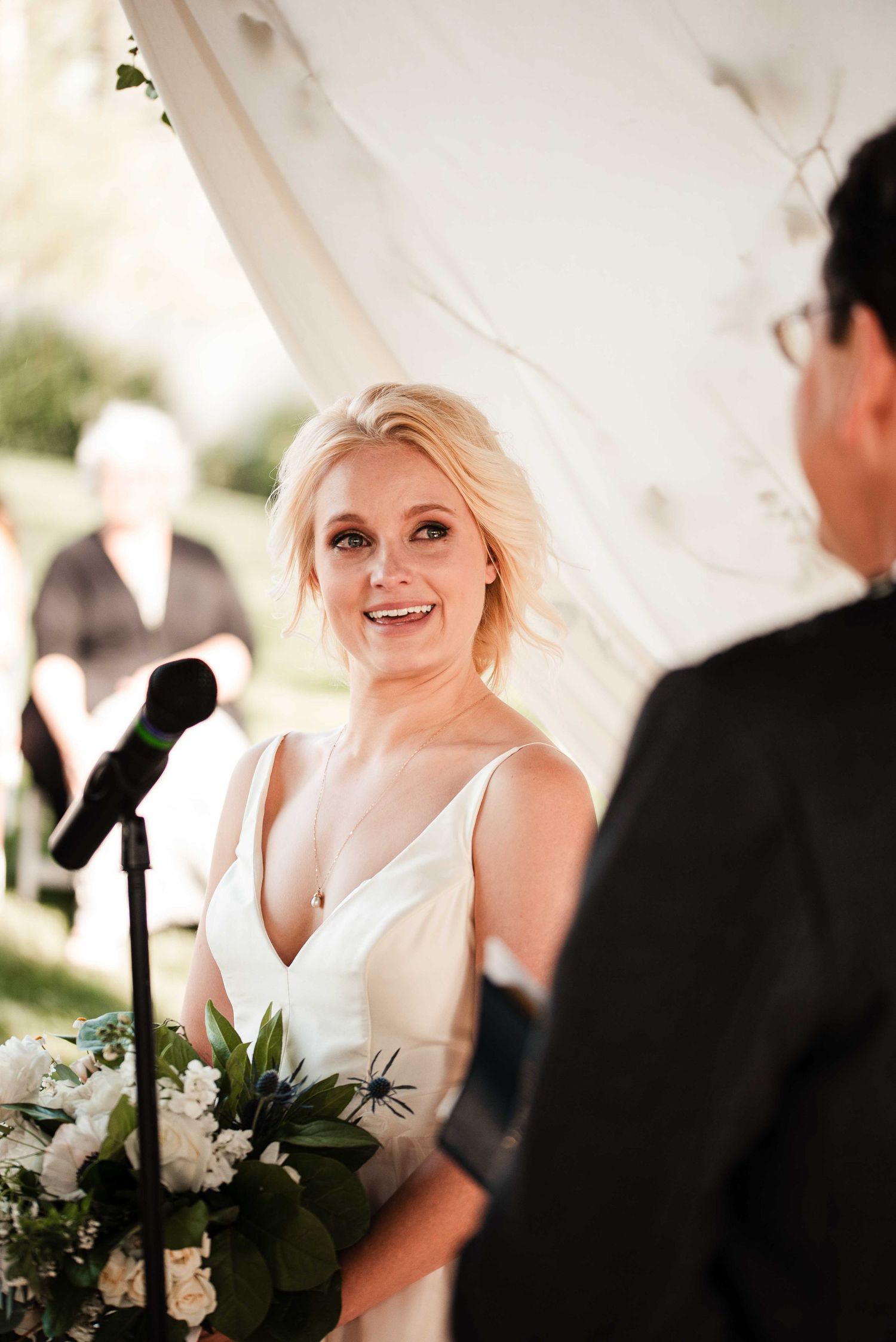 Smiling blonde bride in white gown holding white floral bouquet during outdoor wedding ceremony under tent.