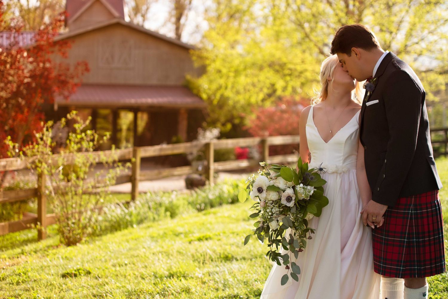 Bride and groom kissing outdoors, she holds a lush bouquet, he wears a red tartan kilt, with a rustic barn backdrop.