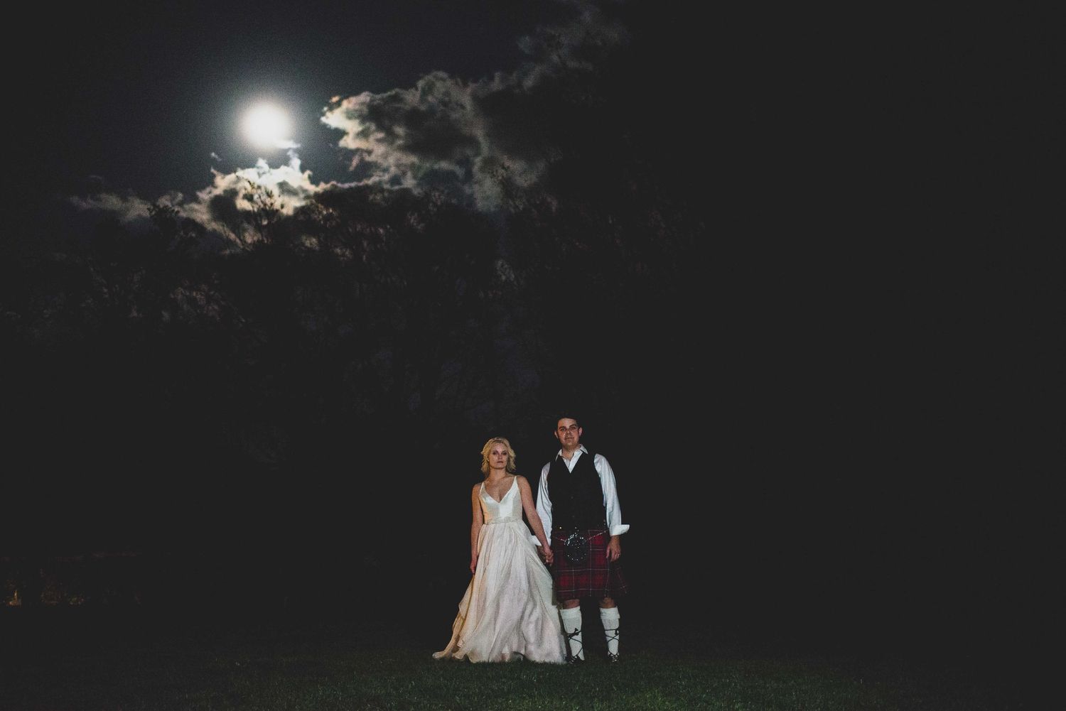Bride in white gown and groom in Scottish kilt stand together under a full moon at night.