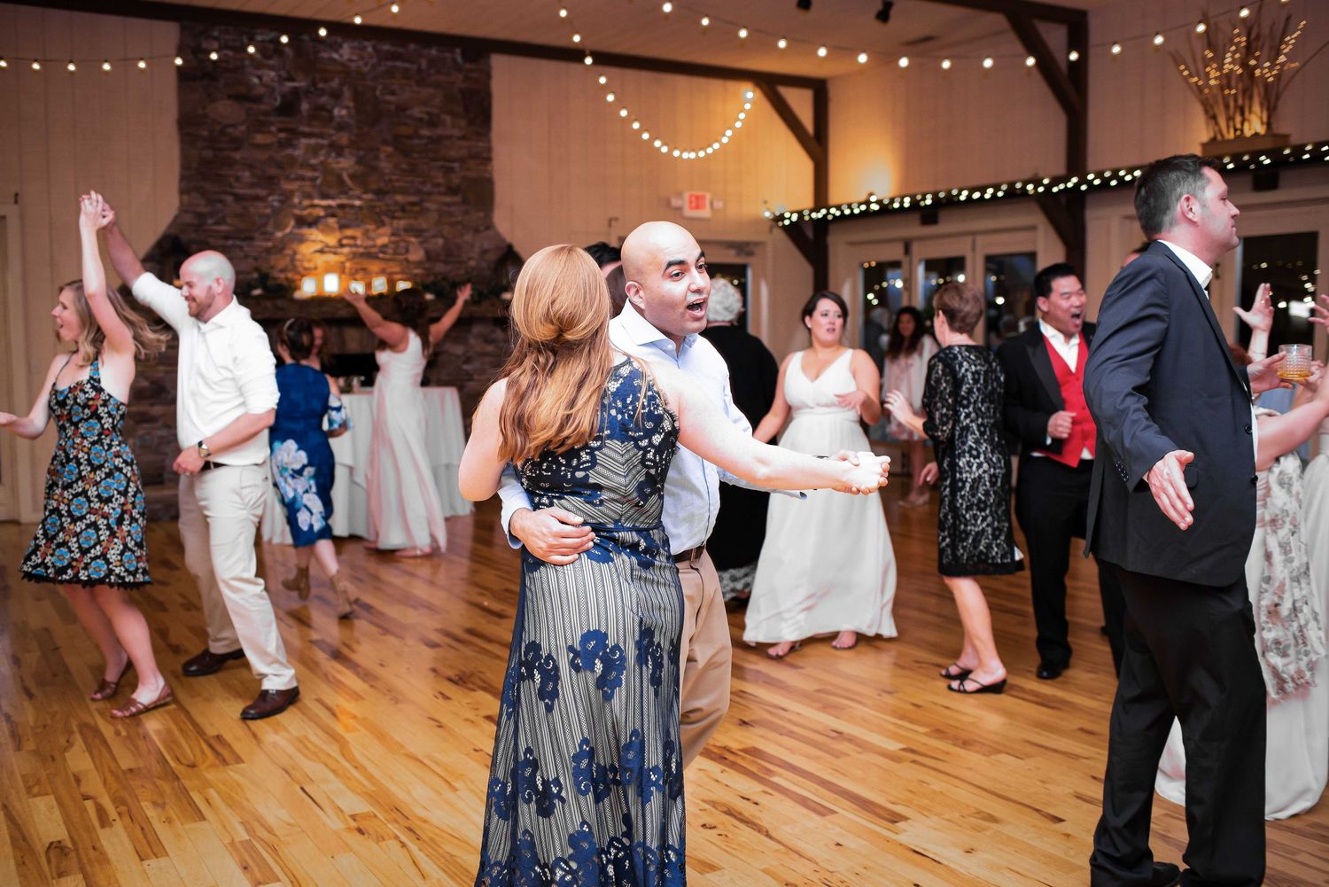 Guests dancing at a rustic wedding reception on a hardwood floor with string lights overhead.