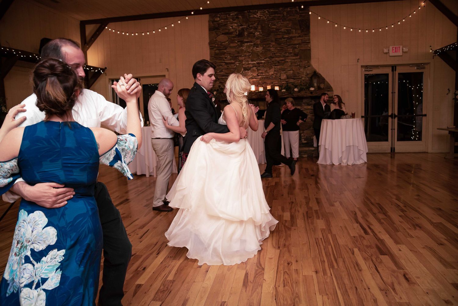 Bride in white gown dances with groom at elegant wedding reception on hardwood floor with string lights.