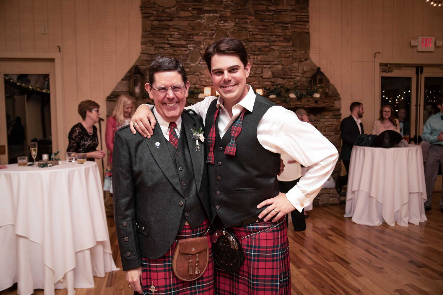 Two men wearing traditional Scottish kilts in red tartan smile together at a formal wedding reception venue.