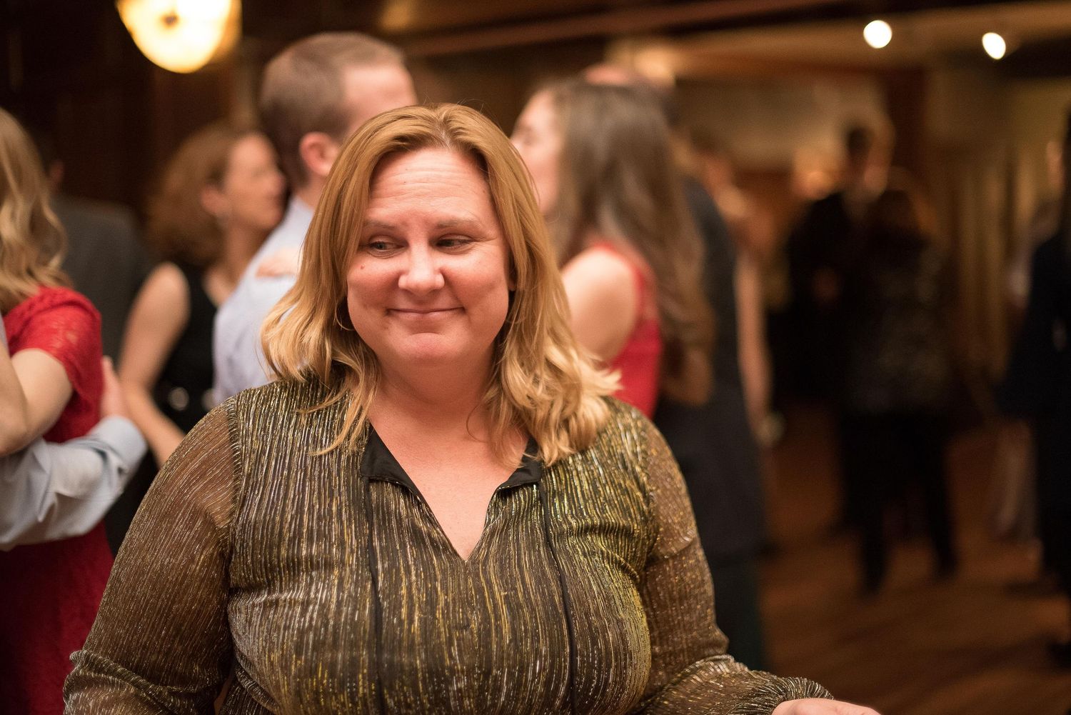 A smiling woman with blonde hair poses at an indoor social event with people mingling in the background.