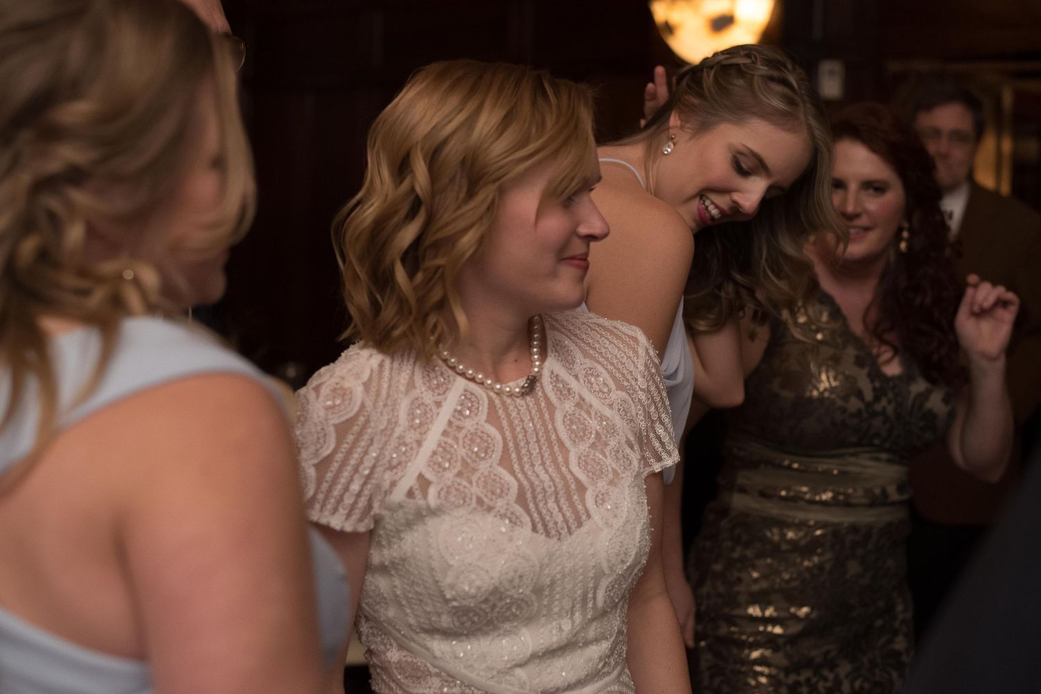 Women in elegant dresses dancing and laughing together at a wedding reception in a warmly lit venue.