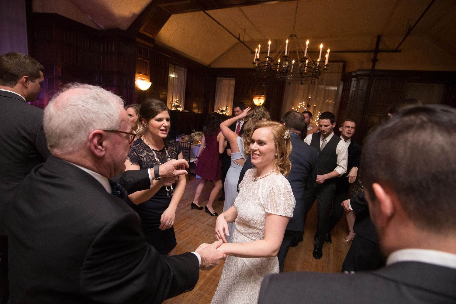 Bride in white dress dancing joyfully with guests at elegant wedding reception venue with chandeliers.