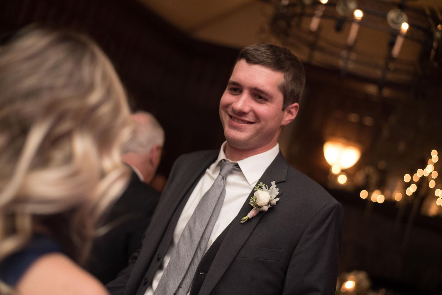 Young man in suit with boutonniere smiling at wedding reception in elegant venue.