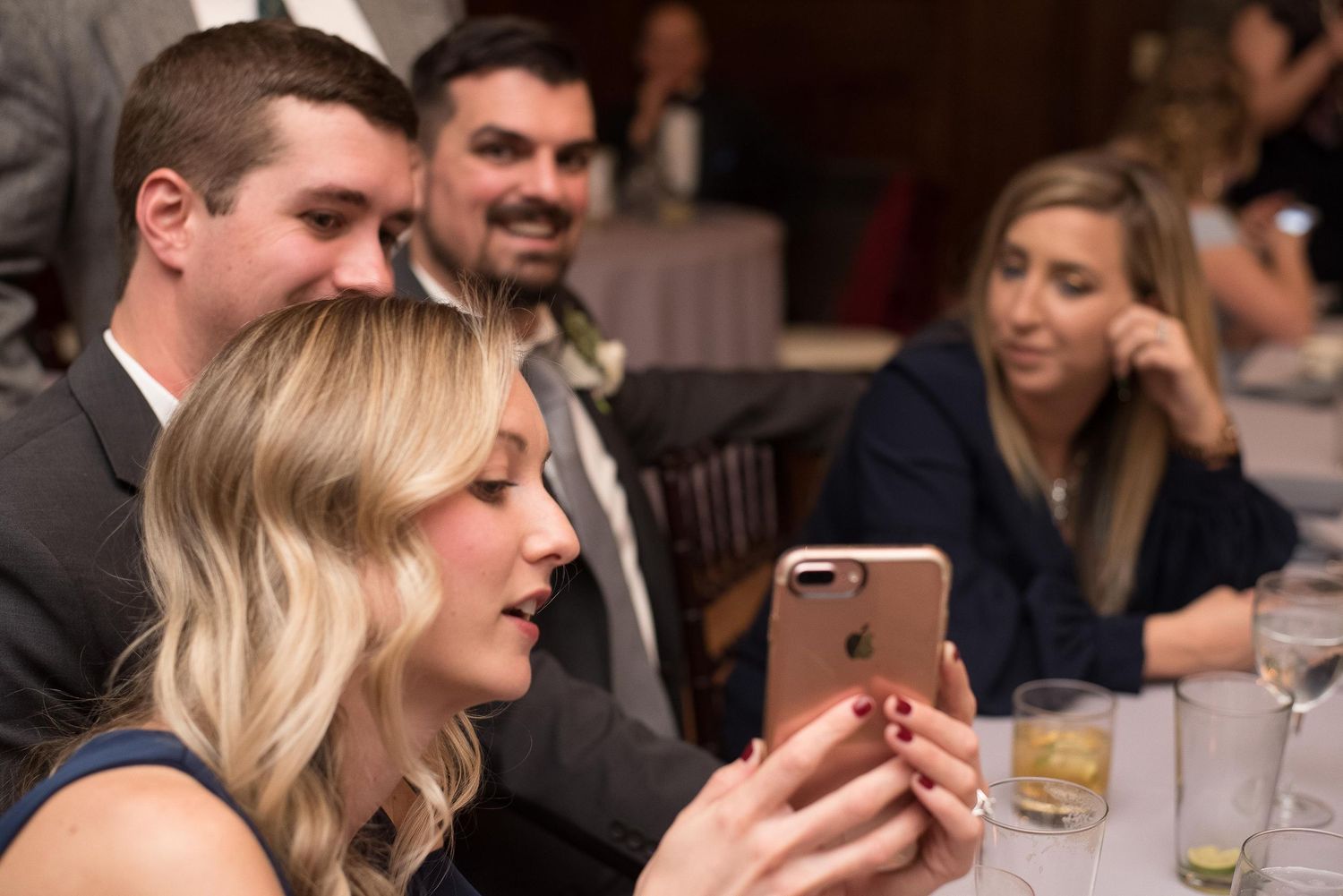 Woman in formal attire views phone at elegant event, guests socializing in background.