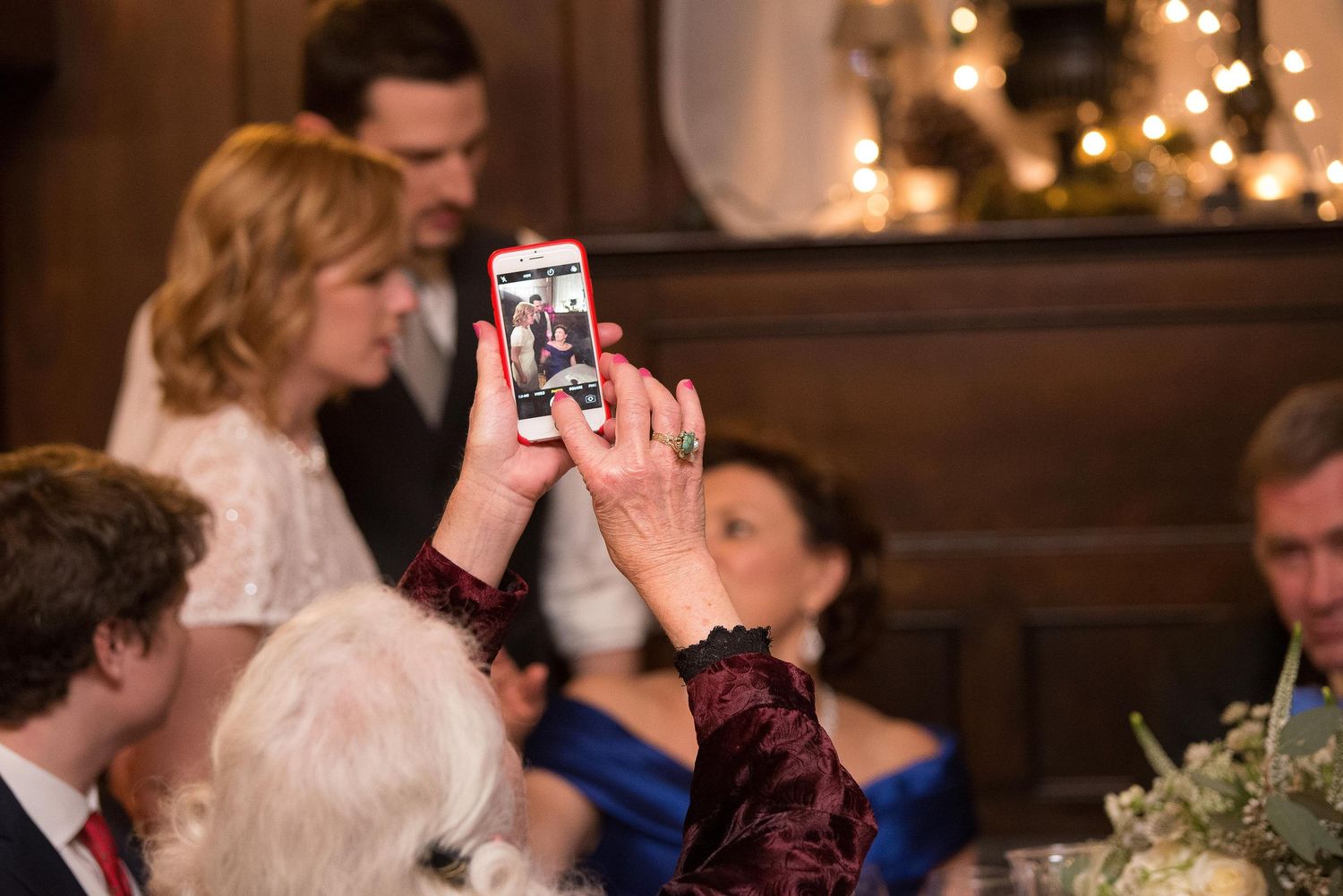 Guest captures bride and groom on smartphone during elegant wedding reception with warm string lights.