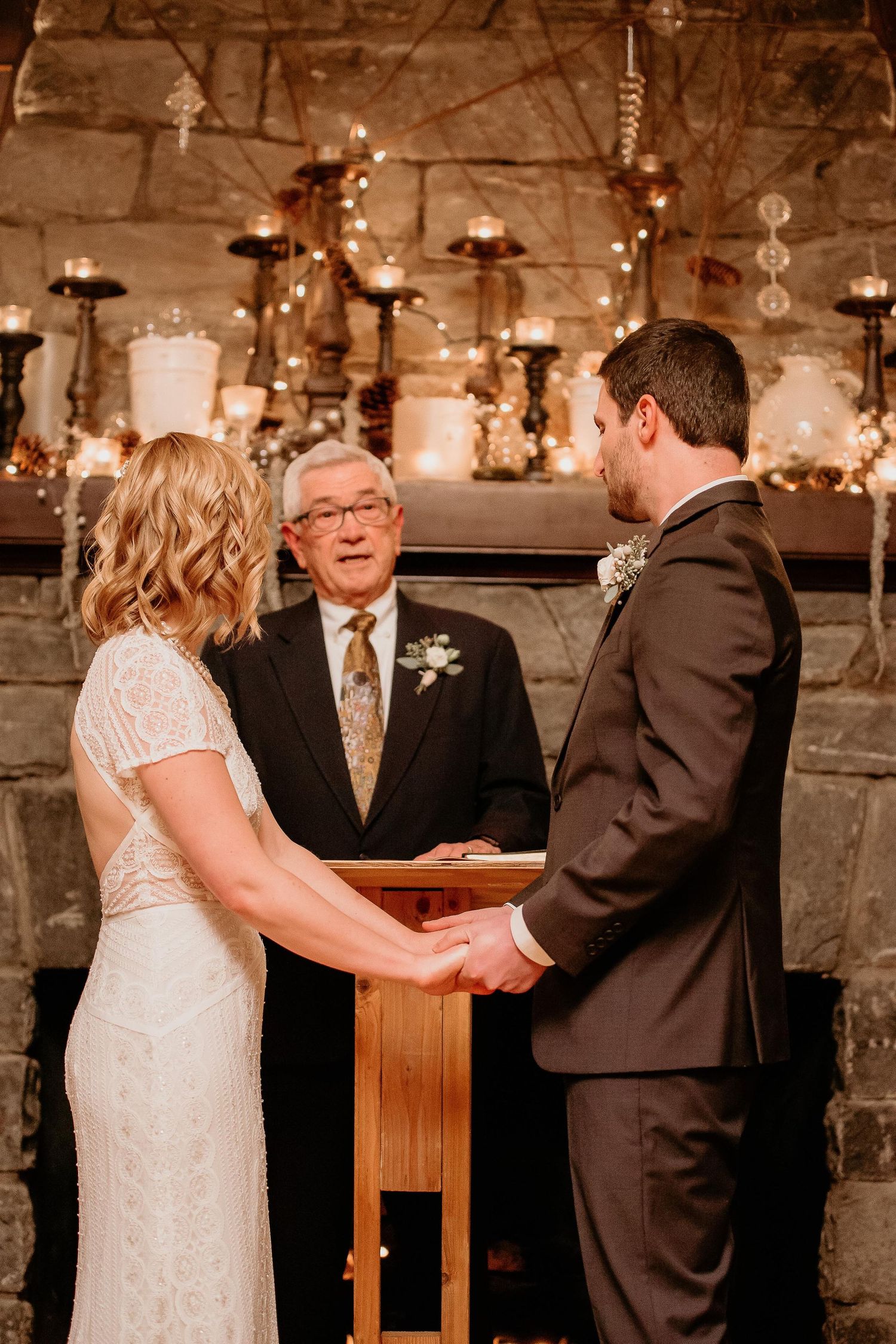 Bride and groom hold hands during wedding ceremony before officiant at candlelit stone fireplace altar.