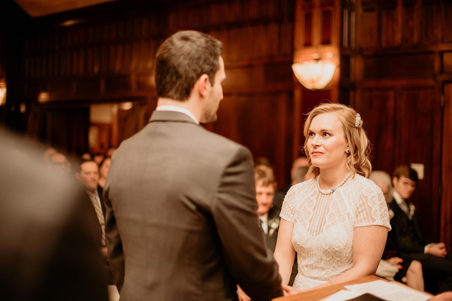 Bride and groom exchange vows at elegant wood-paneled wedding ceremony with guests seated in background.