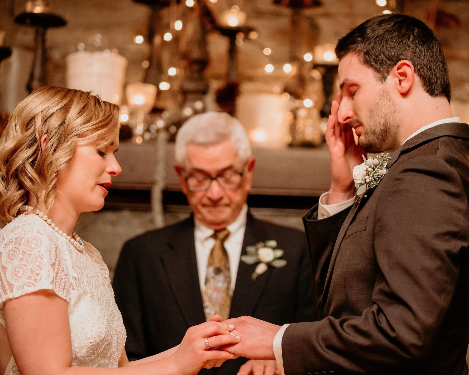 Emotional groom wiping tears during wedding ceremony while bride places ring on his finger.