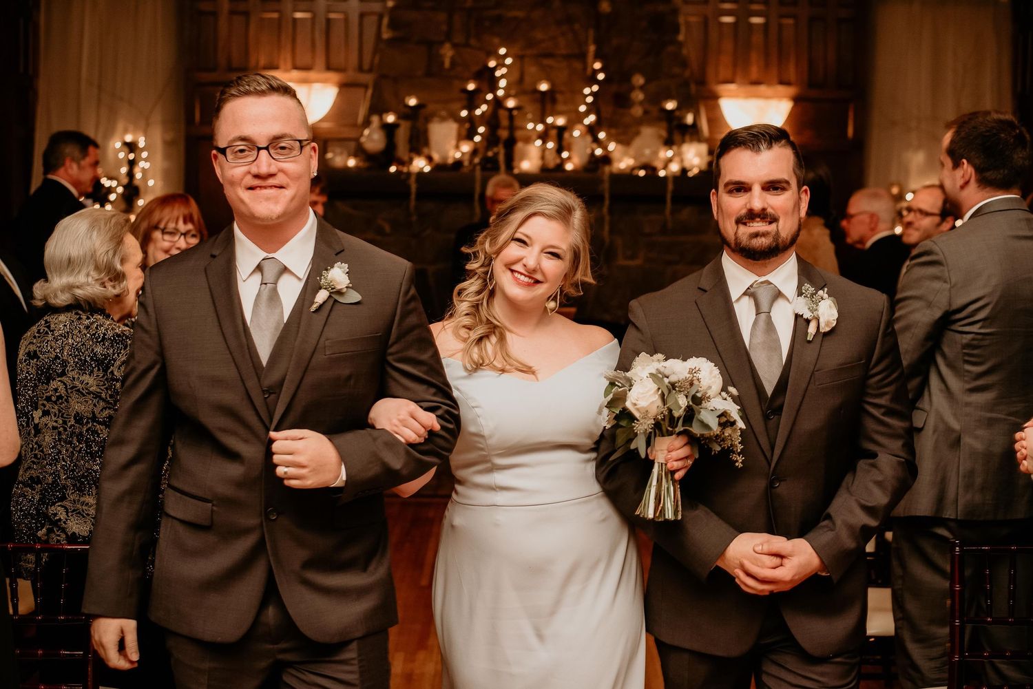 Bride in silver gown walks down aisle with two groomsmen in gray suits at elegant wedding reception.