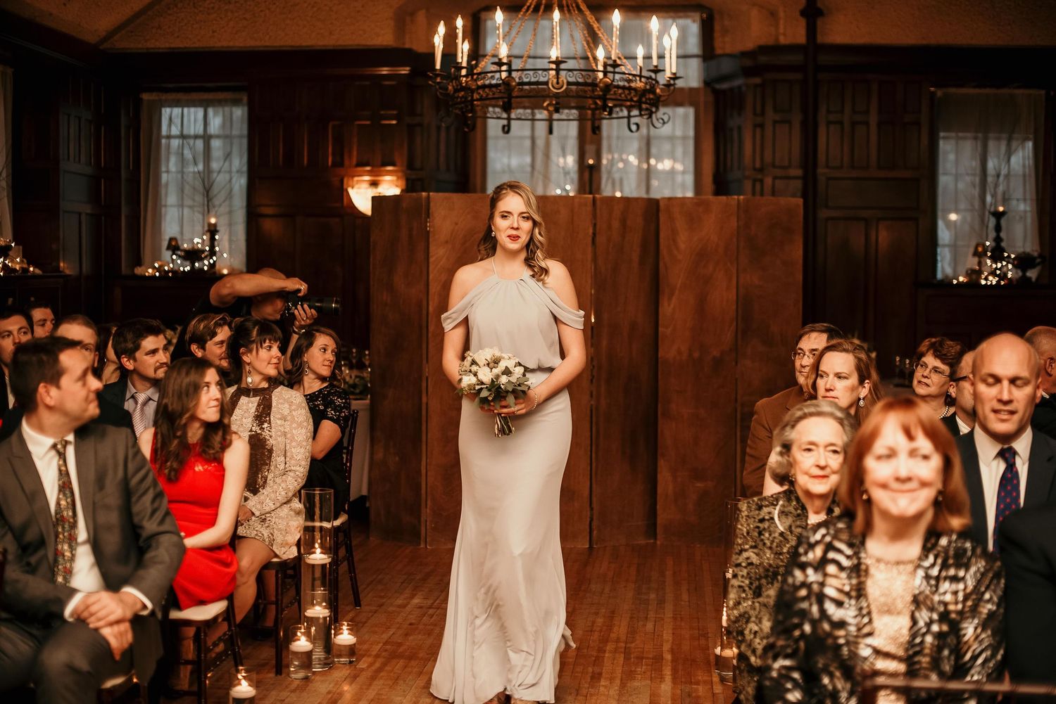 Bridesmaid in silver gown walks down aisle in elegant wood-paneled wedding venue with chandelier.