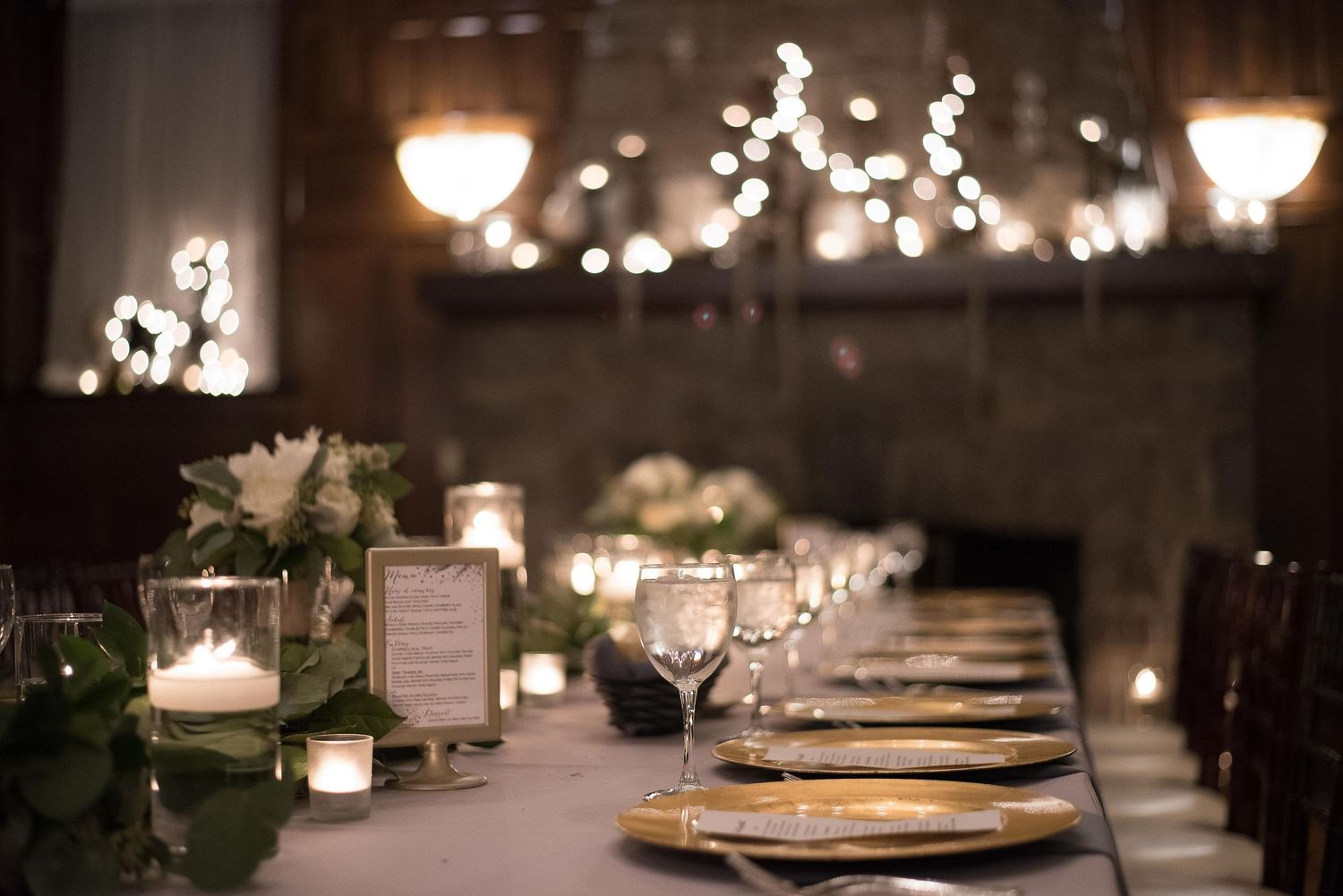 Elegant candlelit wedding reception table with gold chargers, crystal glasses, white floral centerpieces, and soft bokeh lights.