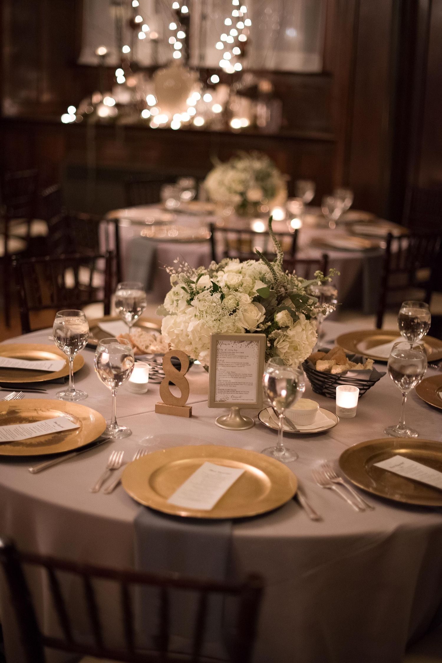 Elegant wedding reception table with gold charger plates, white floral centerpiece, candles, and soft string lights in background.