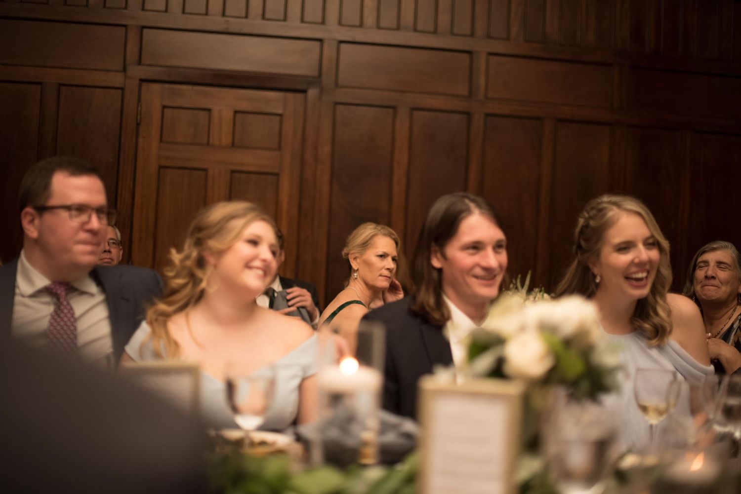 Guests laughing and smiling at a wedding reception dinner in an elegant wood-paneled venue.