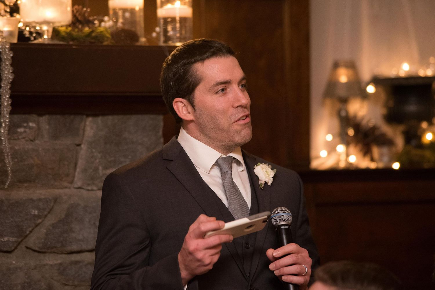 Man in dark suit giving a speech at a wedding reception, holding microphone and notes near candlelit fireplace.
