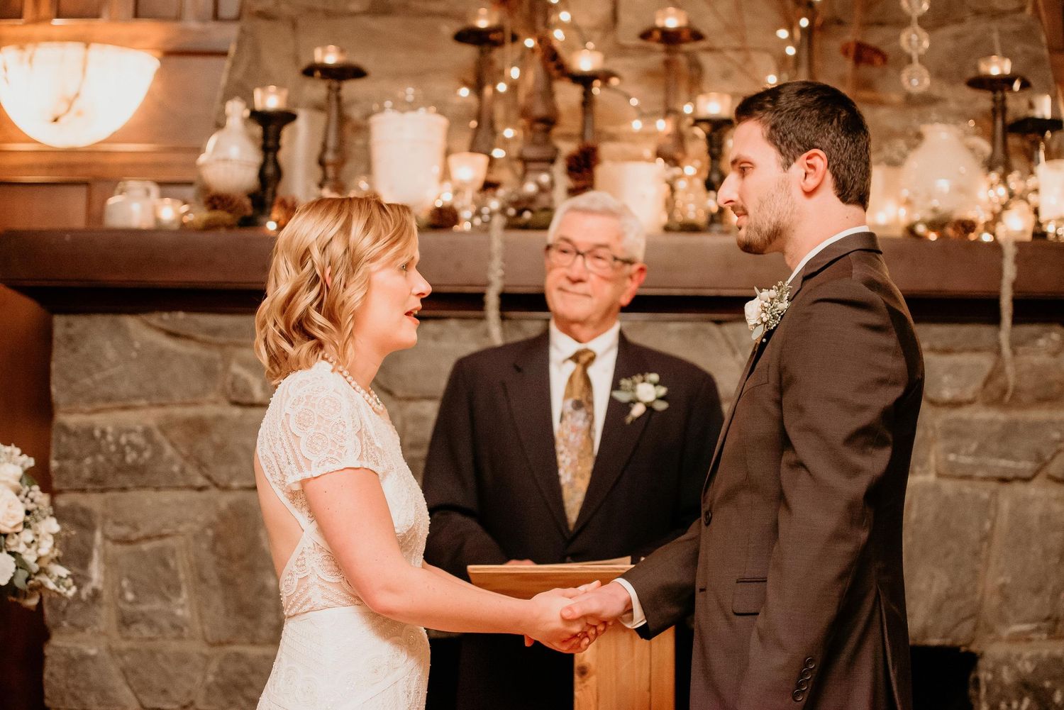 Bride and groom exchange vows holding hands before officiant at rustic stone fireplace wedding ceremony.