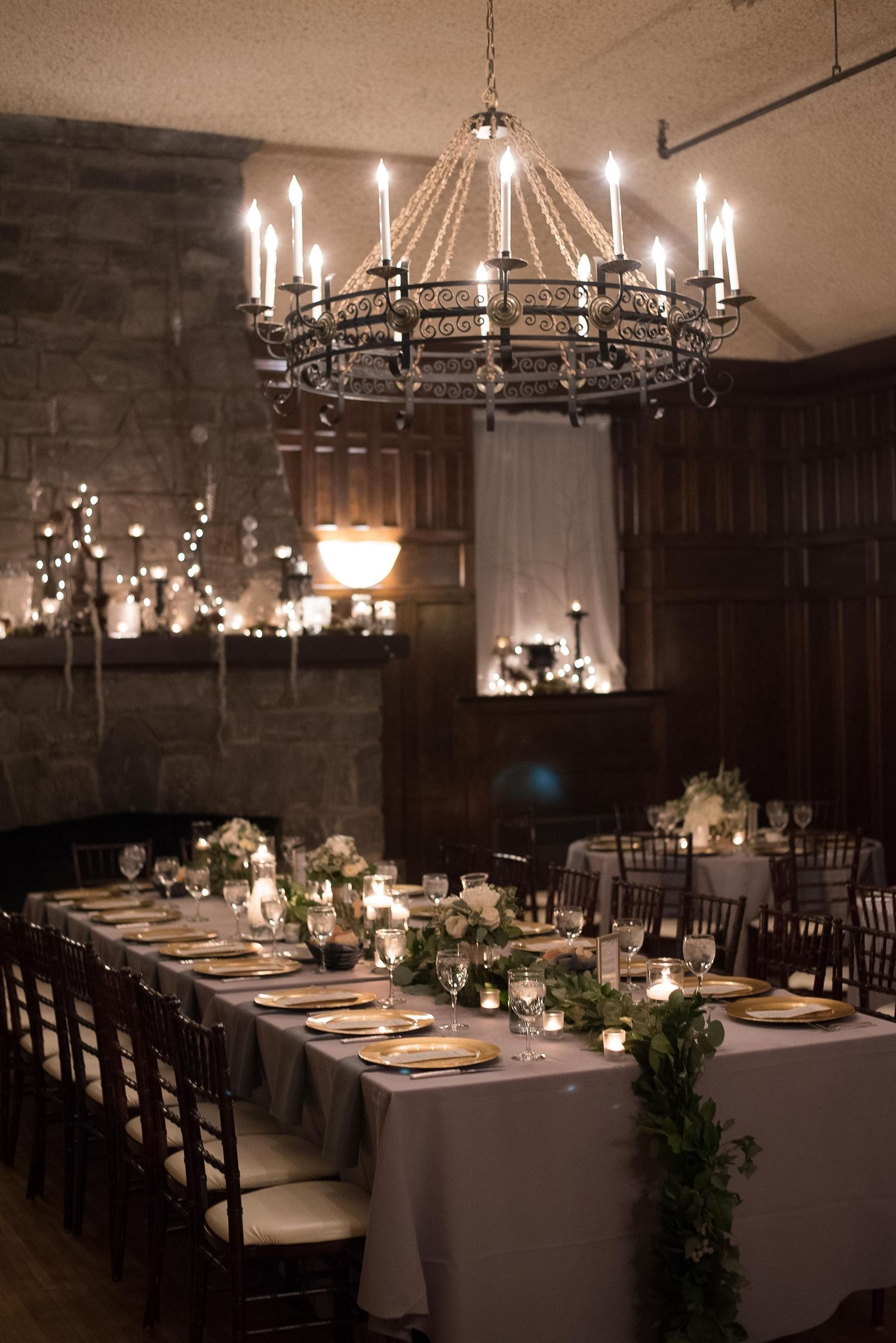 Elegant wedding reception with chandelier, candlelit long tables, greenery runners, gold chargers, and stone fireplace backdrop.
