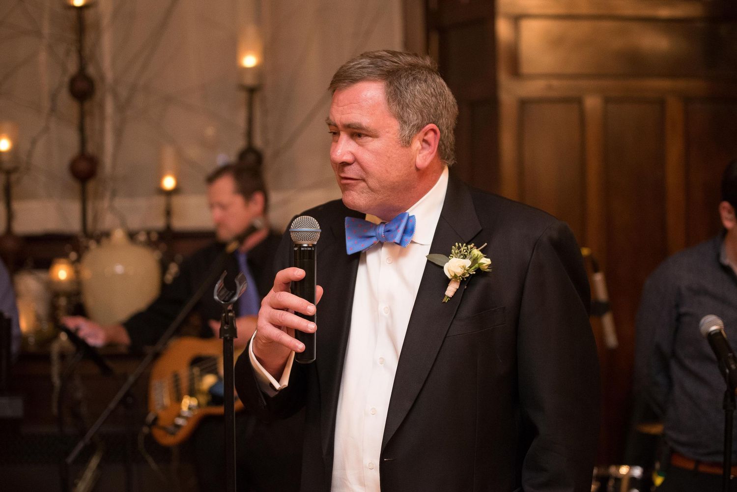 Man in tuxedo with blue bow tie speaks into microphone at formal wedding reception event.