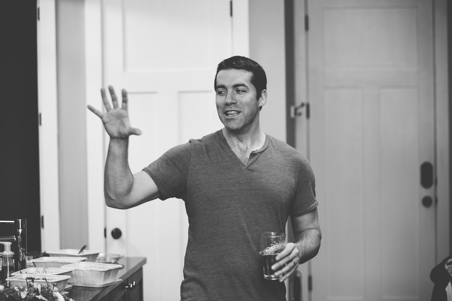 Black and white photo of a smiling man waving while holding a drink at an indoor gathering.