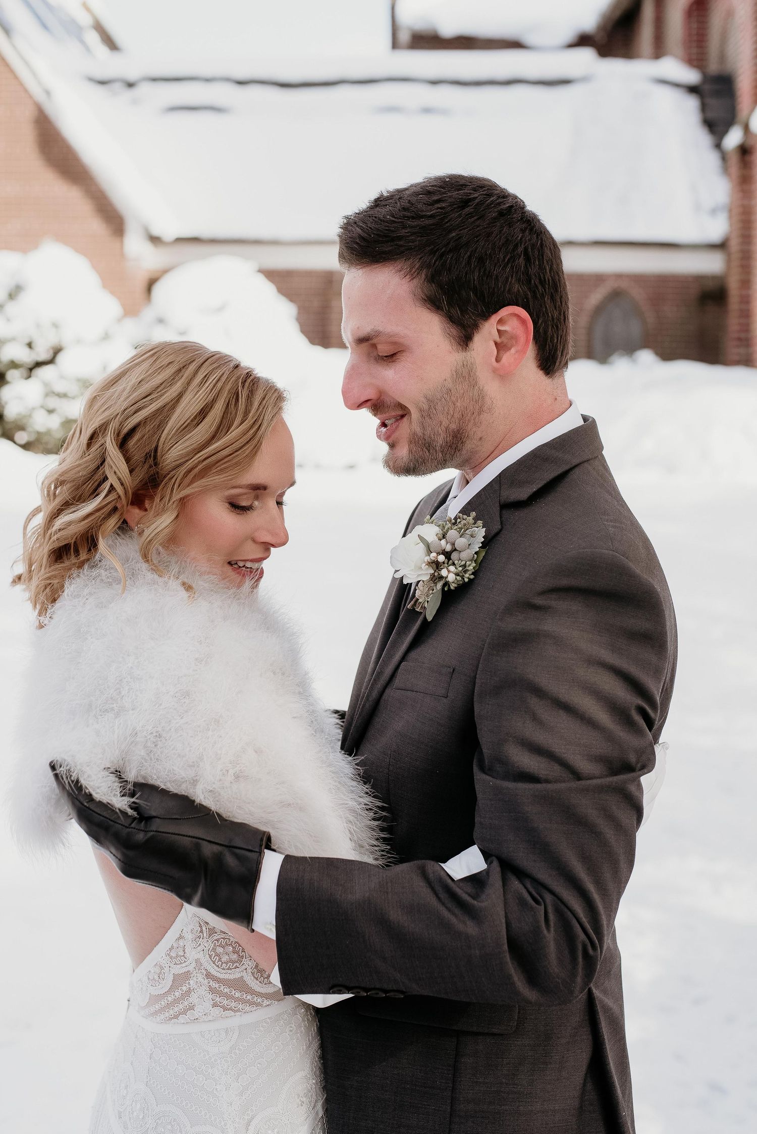 Bride in white lace gown with fur stole and groom in gray suit embrace outdoors in winter snow setting.