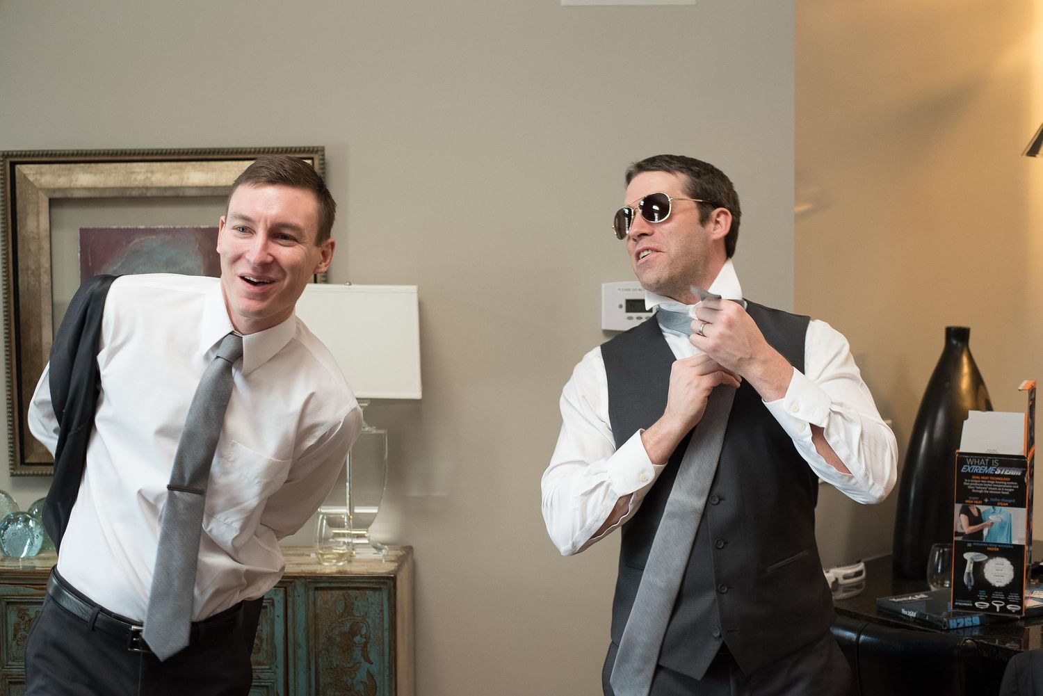 Two groomsmen laughing and adjusting ties while getting ready for a wedding ceremony indoors.