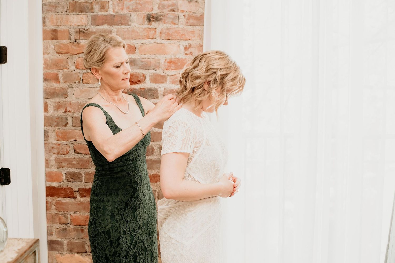 A woman in a green lace dress helps a bride button up her white wedding gown near a brick wall.