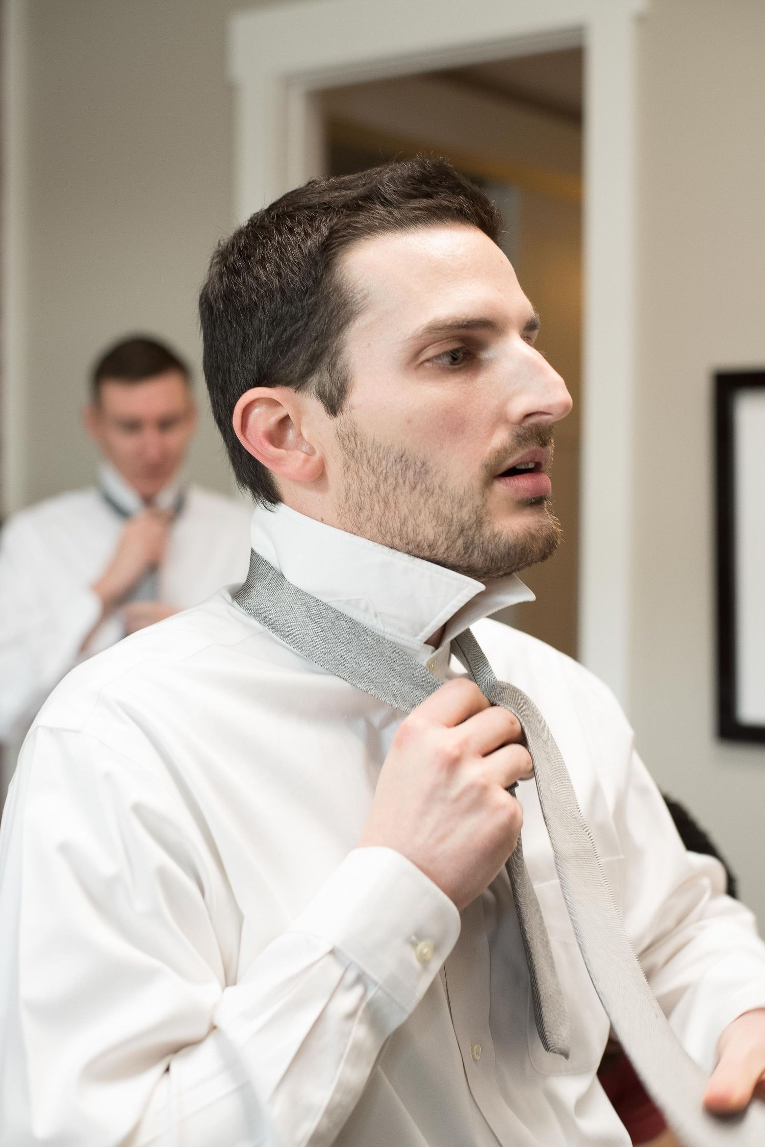Man in white dress shirt adjusting gray tie, with another man tying his tie visible in background mirror.