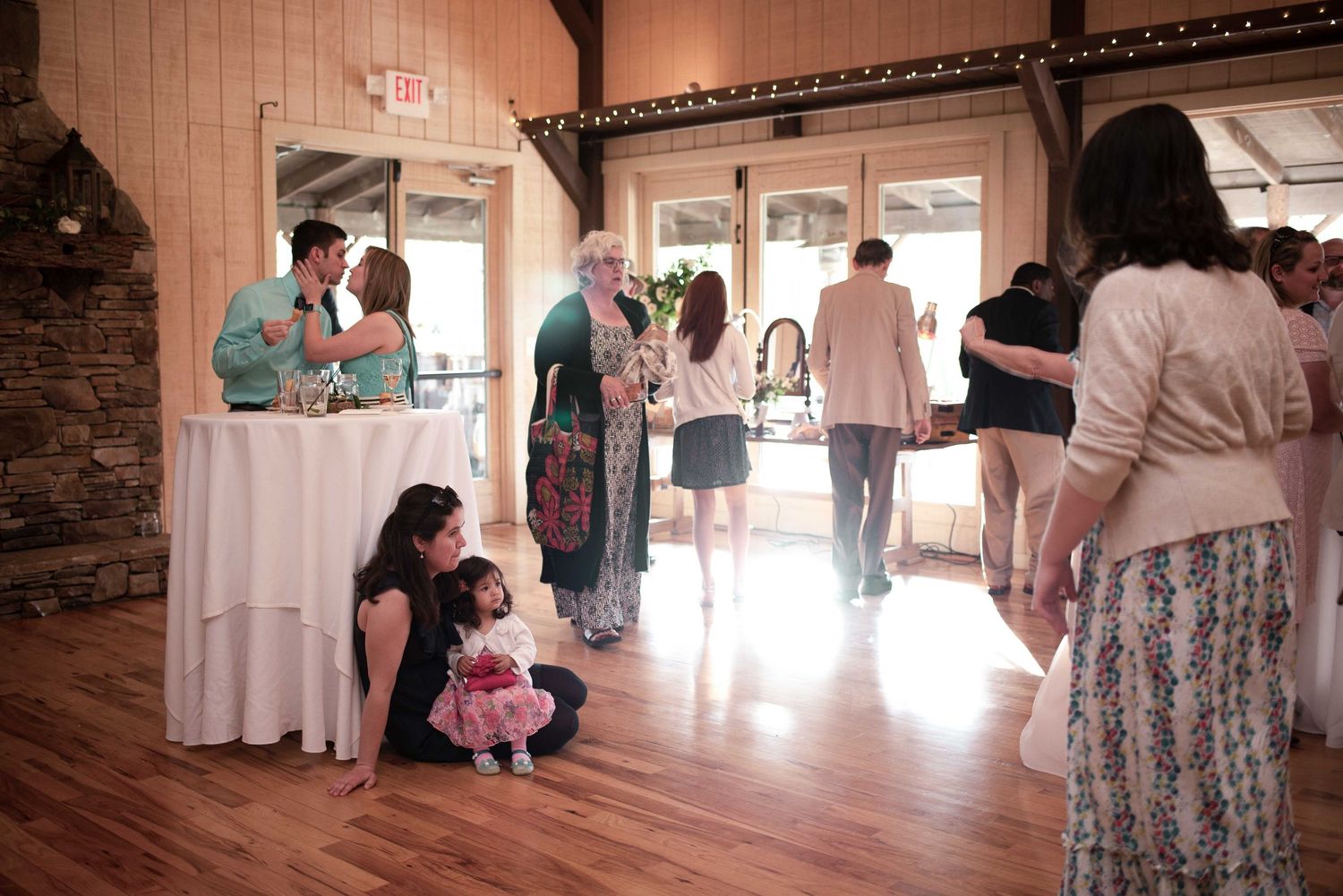 Guests mingling at a rustic wedding reception on hardwood floors, with a child sitting near a white-draped table.