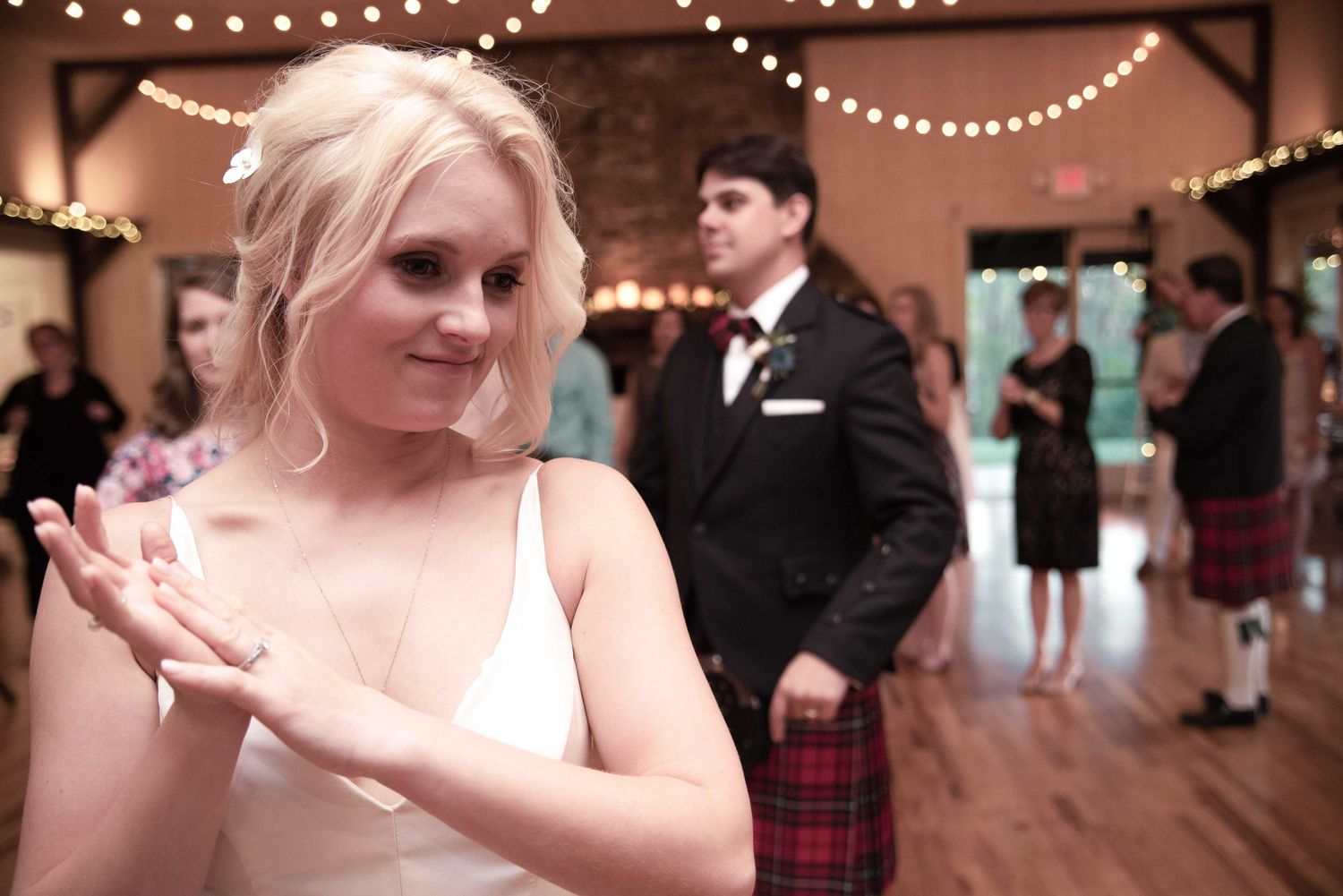 Bride in white dress clapping at wedding reception with groom in kilt and tuxedo in background.